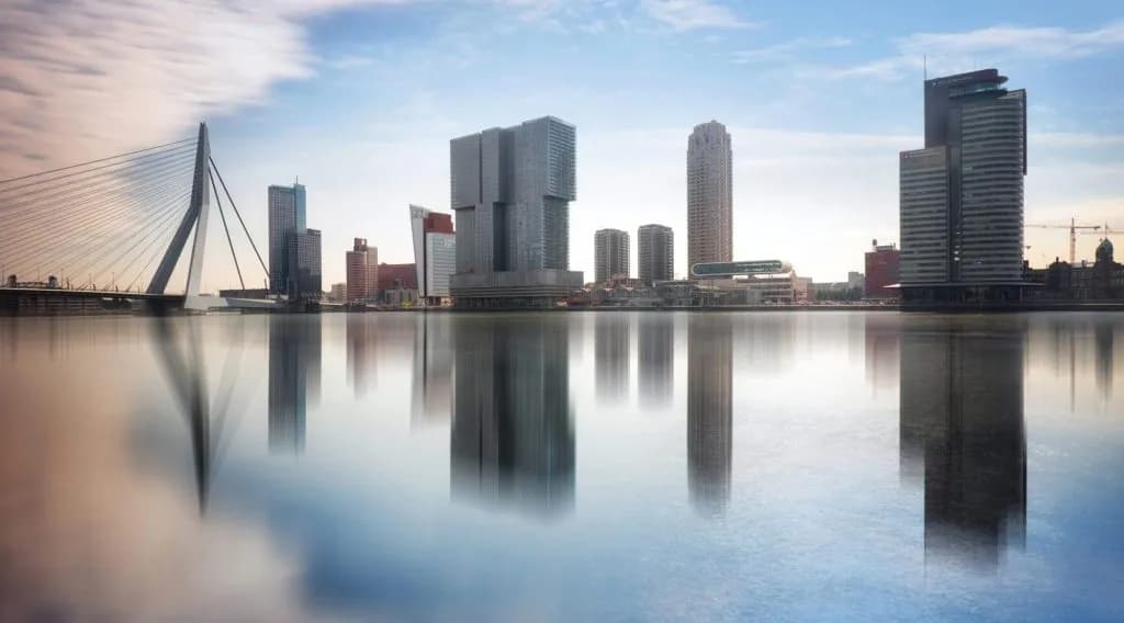 Rotterdam skyline with Erasmus Bridge reflected in calm water under a bright sky.