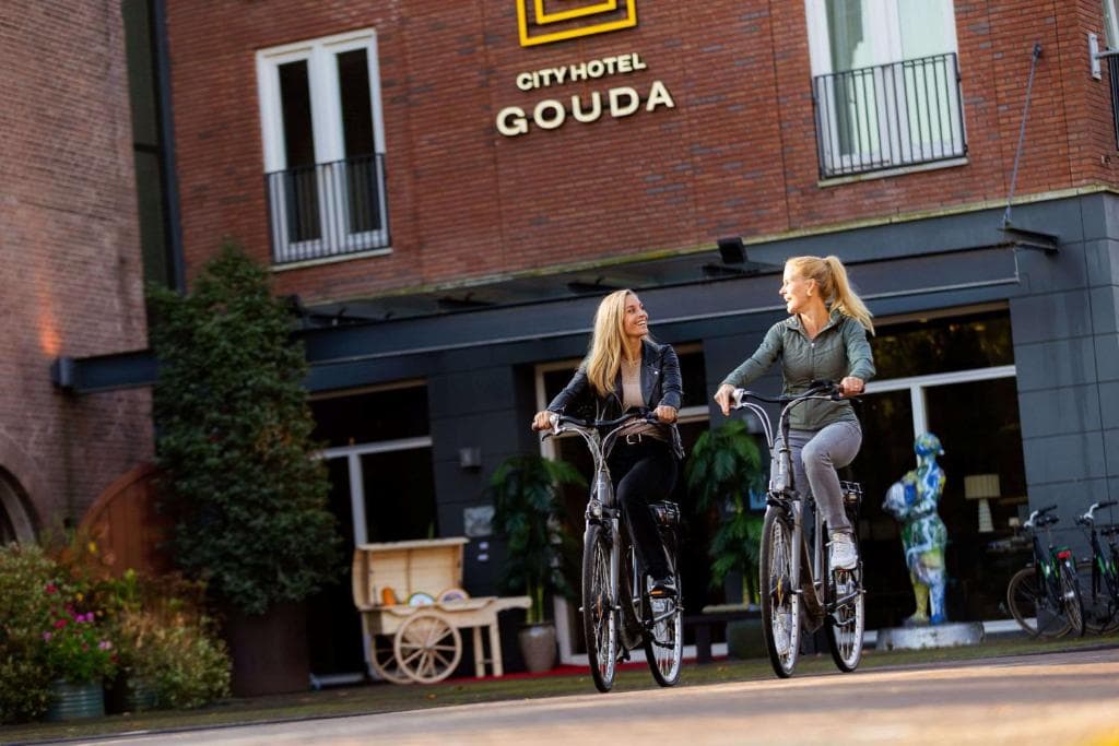 Two women cycling past City Hotel Gouda entrance with decorative cart and statue.